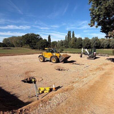 Groundworks for a new farm building, with foundation pads dug into a hardcore base, a telehandler and mini digger working and a laser level set up beside the site