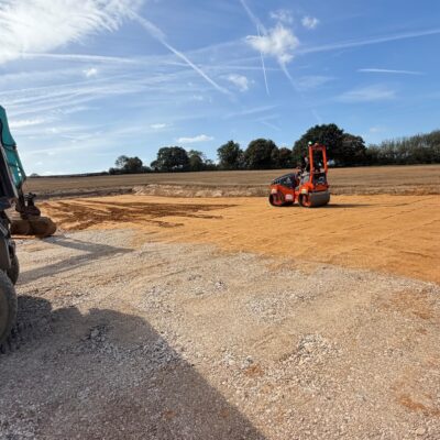 Farm yard base being compacted with a roller on hardcore, with a tipper trailer and digger in the foreground and farmland beyond