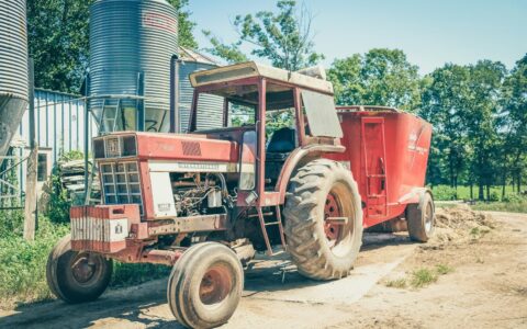 Red tractor and feeder wagon parked on an unsurfaced farmyard beside grain silos