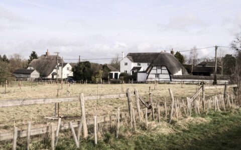 A thatched-roof farmhouse and village cottages bordering an open farm field with post-and-rail fencing, illustrating the type of rural land close to a village that can be released for a rural exception site affordable housing scheme in the English countryside.
